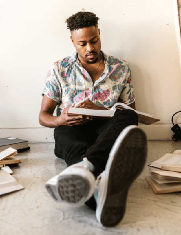 A colored man reading a book on the floor.