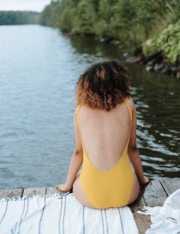 A colored woman with curly hair and yellow one piece sitting on a dock.