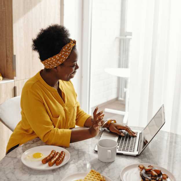 A Colored woman on her laptop while food are on plates on the counter.