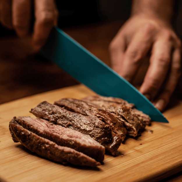 A Colored person cutting up a steak on a cutting board with a blue knife.