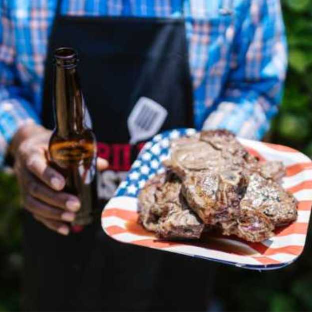 A Colored person holding a plate with steaks in one hand and a beer in the other.