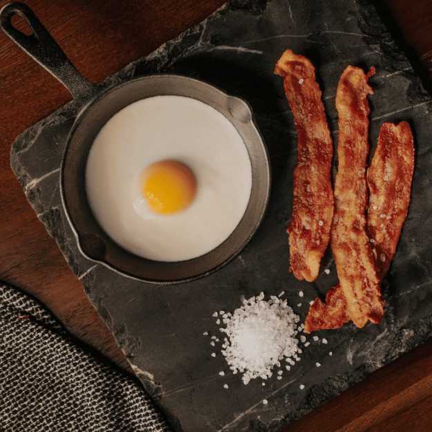 An egg in a pan, salt in a pile, and bacon piled togther.