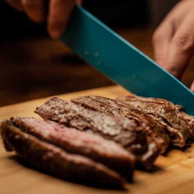Someone slicing meat on a cutting board with a blue knife.