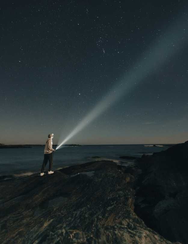 Someone on a sea cliff pointing a flashlight at the stars
