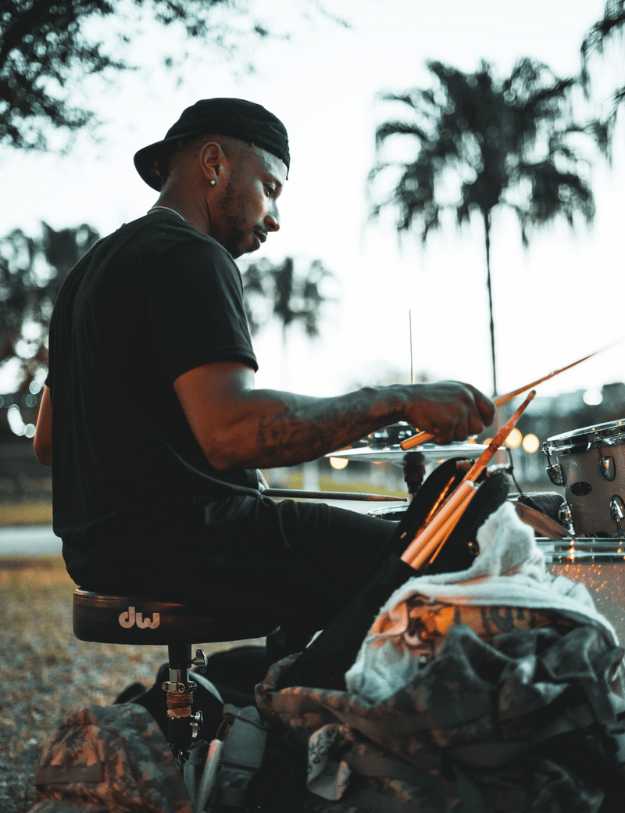 A colored man sitting outside while playing the drums.